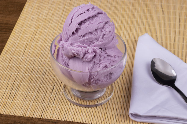 美味的葡萄味紫色冰淇淋装在一个透明的碗里，旁边放着一个勺子，餐巾是白色的。美食摄影冰淇淋店图片下载