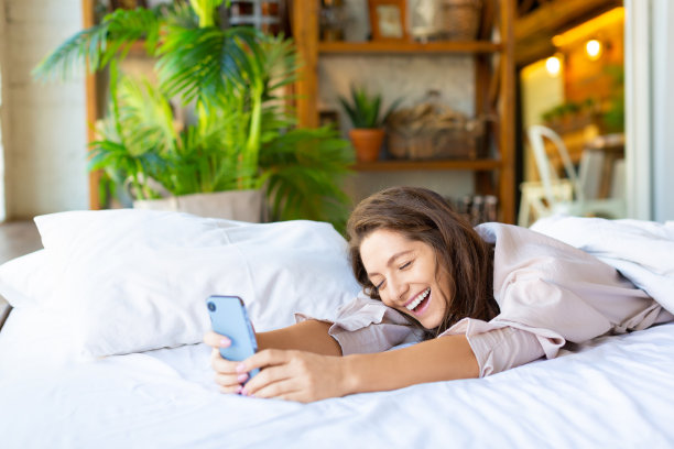 一张快乐大笑的女人在床上拿着智能手机的照片图片下载