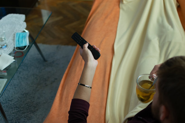 一个生病的年轻人坐在家里，喝着茶，在客厅里休息，吃着药和维生素，量着体温，盖着毯子躺在床上看电视。图片下载