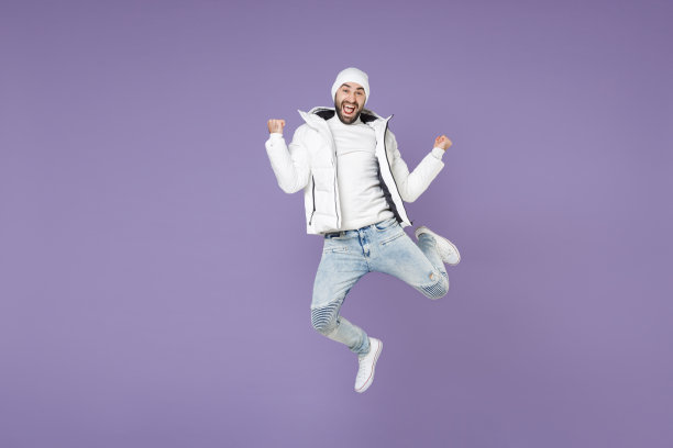 全身快乐的男人穿着暖和的白色棉袄夹克帽子跳着做胜利者的手势庆祝孤立在紫色背景的摄影棚肖像。人们的生活方式寒冷的冬季概念。图片下载