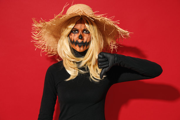 年轻不高兴的女人带着万圣节化妆面具，戴着草帽，黑色稻草人服装，做拇指向下的手势，孤立在纯红色背景上，摄影棚肖像庆祝节日派对的概念。图片下载