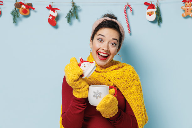 生动的年轻女子，20多岁，戴着黄色的手套，围巾，毛衣，抱开气味，气味，雪人，杯子，咖啡，茶孤立在素色淡蓝色的背景上，画室肖像。2022年新年快乐概念。图片下载