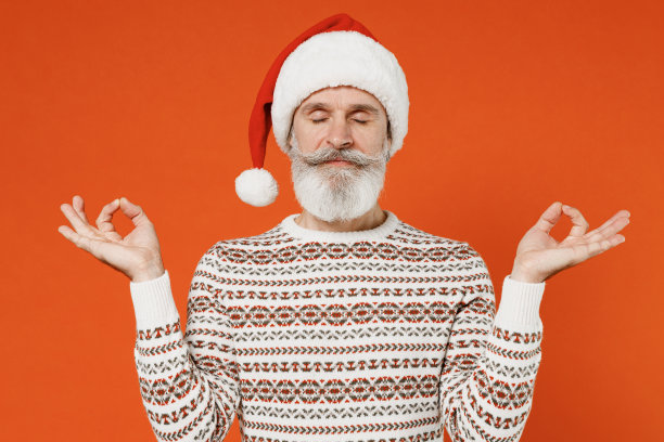 古老的精神宁静的大胡子圣诞老人男人50多岁，戴着圣诞帽，穿着毛衣，摆姿势，握手，展开双手，做瑜伽，冥想，试图平静下来，孤立在素色背景的工作室。图片下载