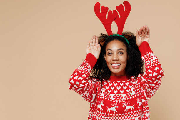 年轻的非洲圣诞老人妇女在圣诞节毛衣装饰鹿角的头做耳朵手势与手孤立在柔和的米色背景工作室肖像。新年圣诞节日概念快乐。图片下载