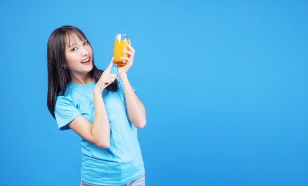 健康水果维生素饮料的概念。享受休闲的年轻女子穿着休闲的指向玻璃与新鲜的有机橙汁在自己的手站立感觉放松在隔离的蓝色背景。图片下载
