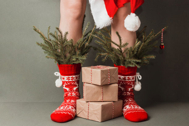 女性腿在红色羊毛袜与装饰品，冷杉树枝和圣诞礼物的绿色背景。圣诞或新年假期快乐，创意概念图片下载