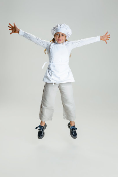 摄影棚里，可爱的小女孩穿着白色的厨师制服，戴着巨大的厨师帽，在白色的摄影棚里孤立地跳着。图片下载