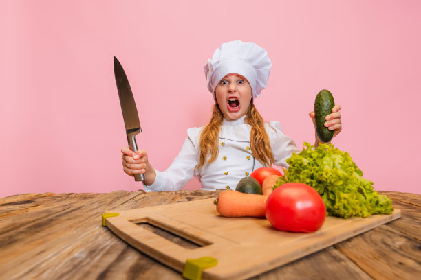 年轻的小女孩穿着白色的厨师制服，戴着巨大的厨师帽，在粉红色的工作室背景下切割新鲜的蔬菜。图片下载