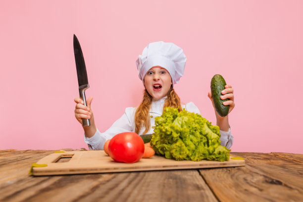年轻的小女孩穿着白色的厨师制服，戴着巨大的厨师帽，在粉红色的工作室背景下切割新鲜的蔬菜。图片下载