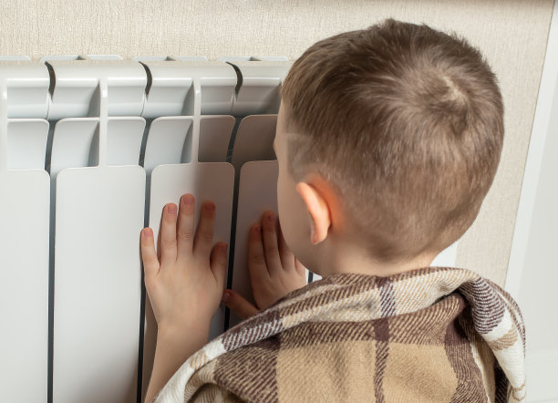一个小男孩在家里靠暖气片取暖。供暖季节的开始。节约热量的概念。能源危机图片下载