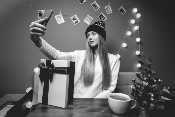 室内女性圣诞肖像。一个女人用手机打视频电话和自拍。冠状病毒和社交距离概念。新年黑白照片与圣诞树和礼盒图片下载