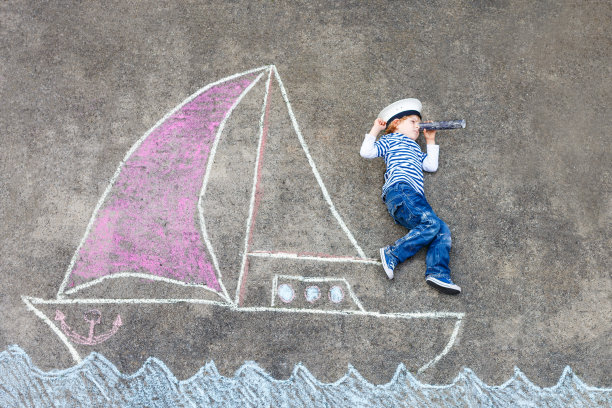 学前班的小男孩在船上或帆船上用彩色粉笔在沥青上画画。夏季儿童户外创意休闲活动。戴着船长帽和望远镜的孩子。图片下载