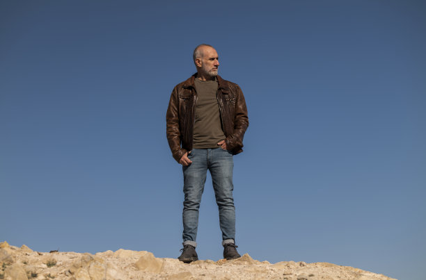 穿着棕色皮夹克的成年男子站在岩石上，在蔚蓝的天空下图片下载