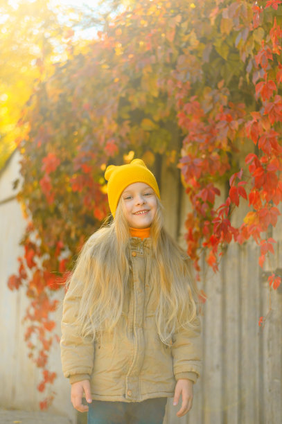 有着金色长发的美丽学前班小女孩微笑着看着镜头，背景是一堵墙，墙上缠绕着野葡萄和黄色、红色和橙色的秋叶图片下载