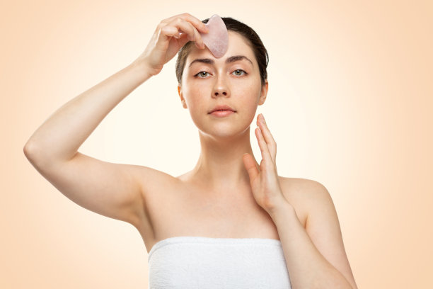 刮痧按摩。一位身穿白色浴巾的年轻女子用石英刮刀按摩额头。米色背景。替代医学和美容的概念图片下载