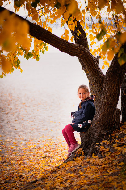 孩子在秋天。小女孩穿着暖和的秋衣，坐在河边，欣赏着清澈的水中和地上美丽的黄叶图片下载