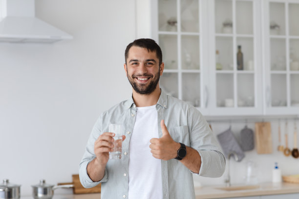满脸胡须的欧洲成年男子面带微笑，手里拿着一杯水，大拇指朝厨房内部图片下载