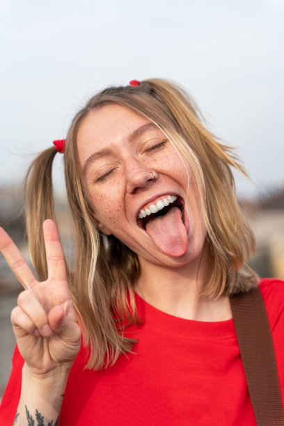 一个年轻的金发女人的竖像，脸上有雀斑，两条滑稽的马尾辫，顽皮地用手指露出舌头和V形手势，正面照片。十几岁的孩子在鬼混。图片下载