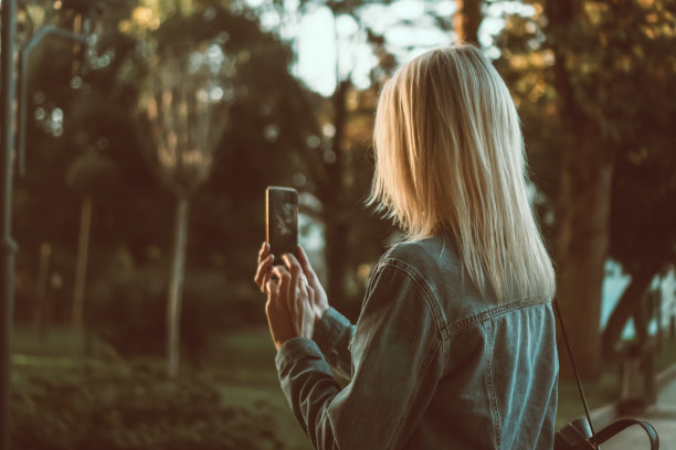 年轻的金发女郎穿着牛仔夹克，用手机拍下公园里美丽的风景，从后面拍照。移动摄影的概念，旅行图片下载