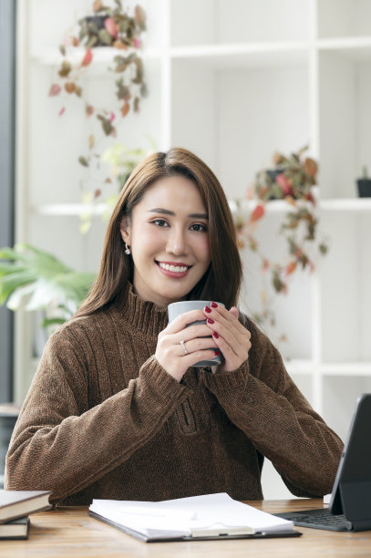 年轻的亚洲女人的肖像穿着羊毛衫，拿着杯子，微笑，看着相机坐在办公桌。图片下载