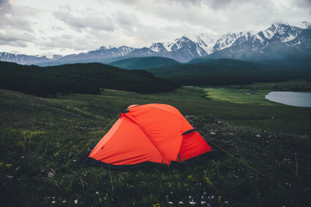 大气的高山景观与橙色帐篷背景的湖和大雪山在阴天。令人惊叹的绿色风景，橙色帐篷，云天下的大山。图片下载