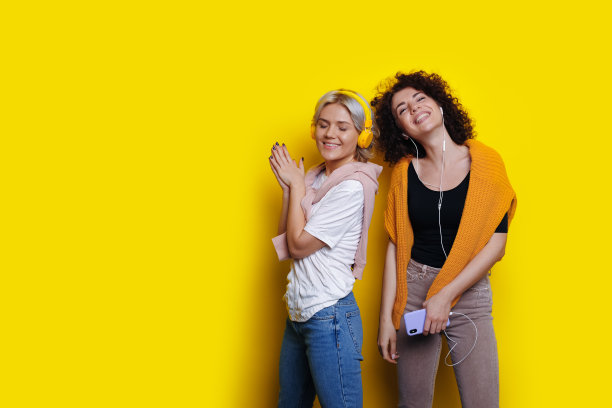 卷曲头发的姐妹们在自由空间的黄色工作室墙上听音乐，戴着耳机对着镜头微笑，使用手机图片下载