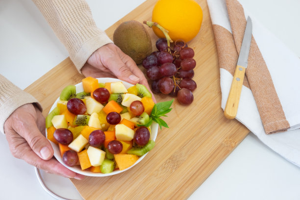 女人们的手捧着一碗五颜六色的新鲜水果。苹果，猕猴桃，橘子，木瓜，芒果，甜瓜。早餐或休息，健康饮食。木制的砧板放在桌子上图片下载
