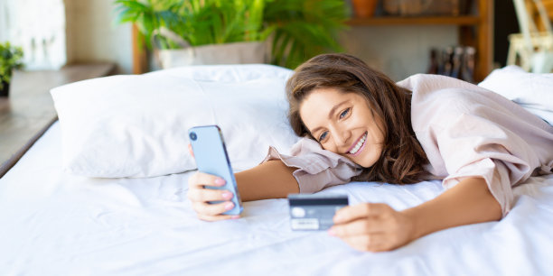 一个开心微笑的女人躺在床上通过手机应用下单。她在网上购物时拨卡号。格式照片2 x1图片下载
