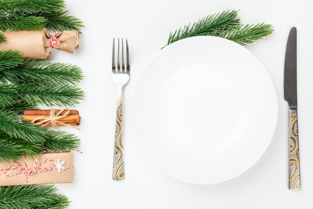 平放在银餐具刀叉旁边白色的空盘子放在白色的桌子上，桌子上有圣诞树上的树枝、礼物和肉桂图片下载