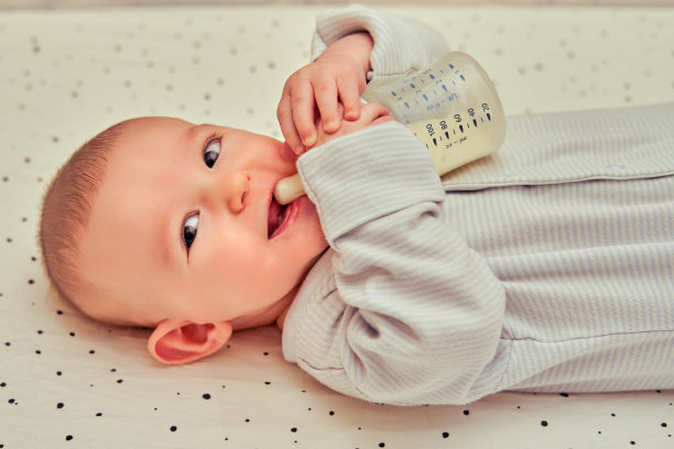 快乐的宝宝手里拿着一瓶牛奶，独自微笑着。三个月大的孩子自己躺在床上图片下载