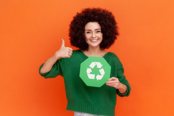 积极的女人，留着非洲式发型，穿着绿色休闲风格的毛衣，露齿的微笑看着镜头，竖起大拇指，举着回收的牌子。图片下载