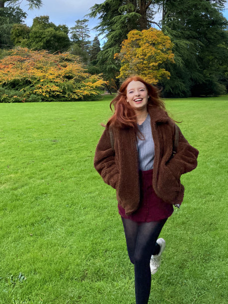 特写照片，有一头红色长发的漂亮女人在公园的草坪上奔跑，标本针叶树包括苏格兰松树，黎巴嫩雪松和喜马拉雅雪松在秋天的阳光下，晴朗的蓝天和蓬松的云图片下载