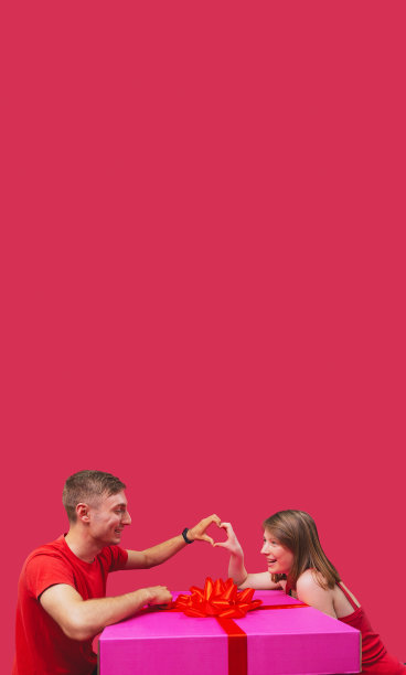 上面的空间照片是两个相爱的人，男女朋友都穿着红色的衣服，坐在大粉色礼物盒旁边，用手指做心形手势。图片下载