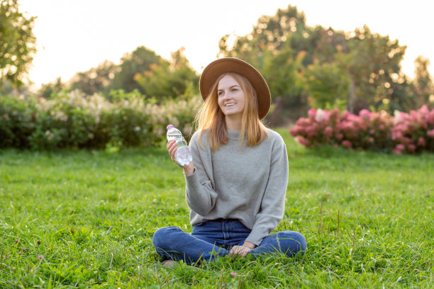 在夏天的绿色公园里，一个戴着帽子的年轻漂亮的金发女人正在喝塑料瓶里的水。食物，休息或生态概念图片下载