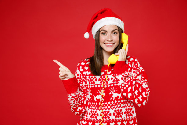 微笑的年轻圣诞老人女人穿着毛衣圣诞帽讲电话，食指指着一边孤立在红色背景的摄影棚肖像。新年庆祝快乐节日概念。图片下载