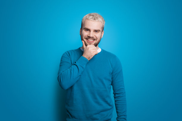 蓝墙上的金发男子正摸着下巴。微笑的人。前视图。英俊的男人。积极的人。图片下载