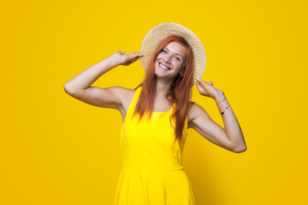 可爱的姜黄色女人正对着摄影棚里的黄色墙壁微笑，手里拿着她漂亮的帽子图片下载