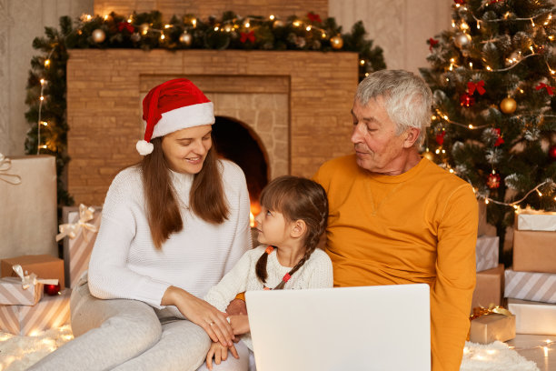 室内照片:一个穿着橙色毛衣的成熟男人和他的家人在充满节日气氛的客厅里，在圣诞树和壁炉旁摆姿势，在打开的笔记本电脑前摆姿势，在线聊天。图片下载