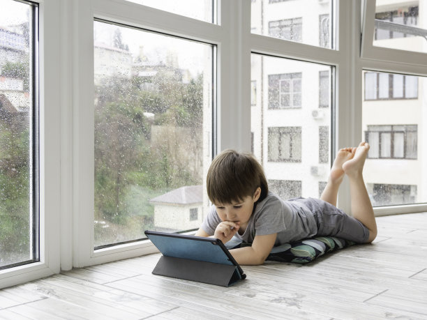 好奇的男孩在电子平板电脑上看卡通片。孩子躺在地板上使用电子设备。外面下雨的时候，孩子们的室内休闲。图片下载