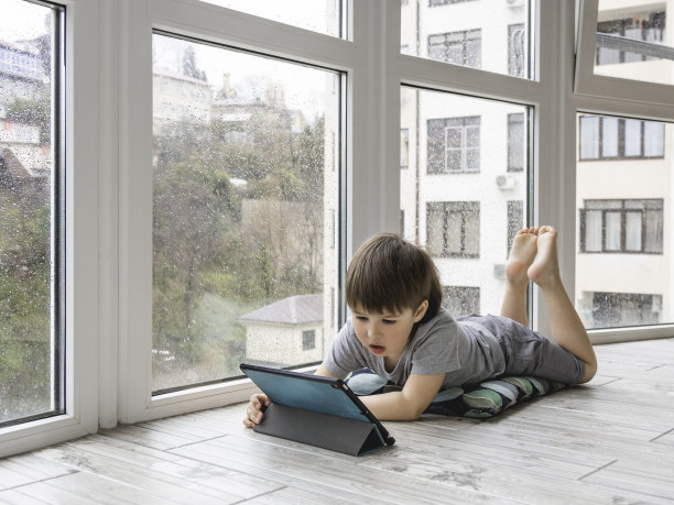 好奇的男孩在电子平板电脑上看卡通片。孩子躺在地板上使用电子设备。外面下雨的时候，孩子们的室内休闲。图片下载