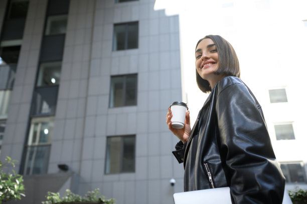 从下面的观点，自信的女人，自由职业者，微笑着看着相机，沿着街道走在现代公司大楼下，手臂下的咖啡外卖笔记本电脑图片下载