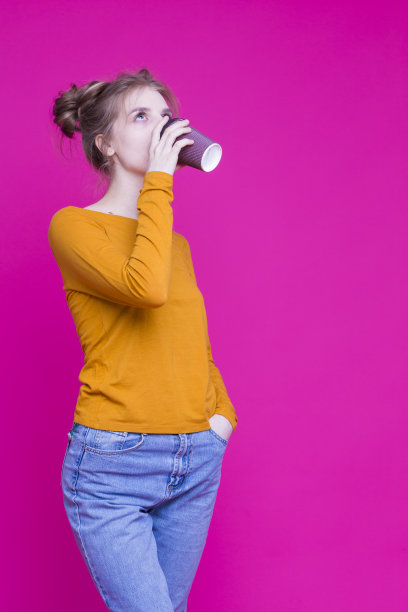 年轻和美丽的白人女孩喝一次性纸杯摆姿势，以无缝的粉红色背景。垂直的图像图片下载