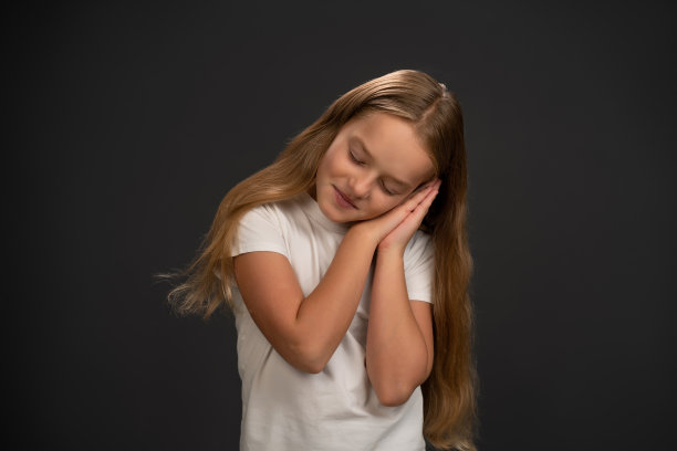 小女孩双手合在一起，头靠在双手上，示意她睡着了。女孩穿着白色t恤对着相机微笑孤立在深灰色或黑色背景图片下载