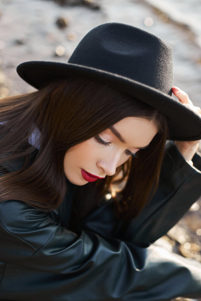 为日落时的年轻女子画像。穿着黑色圆帽和皮革雨衣的时尚女子在湖边。漂亮的妆容和鲜红的唇膏图片下载