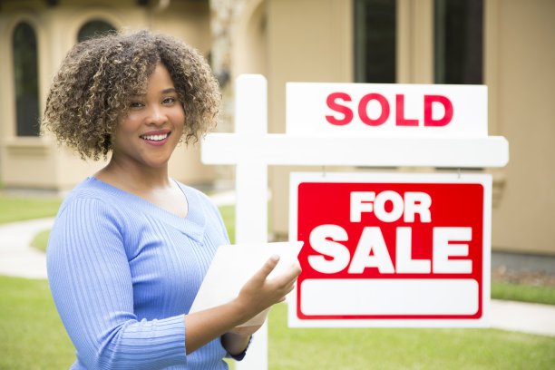 可爱的年轻成人房地产经纪人站在她旁边的出售标志在家门口的院子里。她拿着一个文件夹，穿着蓝色上衣和牛仔裤。图片下载