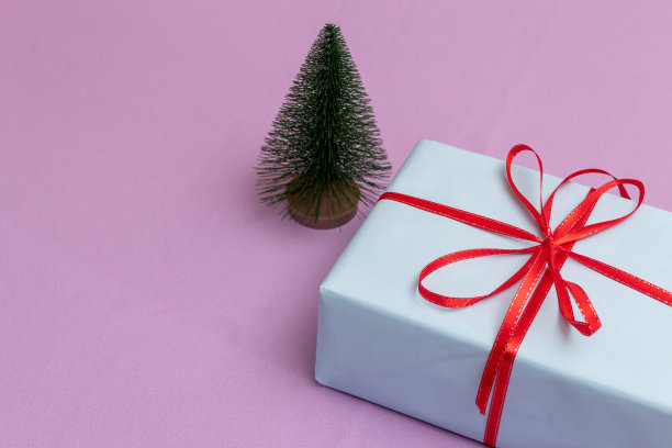 盒子里装着蓝色的纸和红色的蝴蝶结。有一棵小小的人造树。紫色背景与文字的地方。广告的礼物。有选择性的重点。图片下载