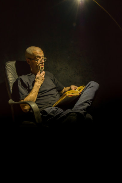 一位老人坐在黑暗中看书，只点着一盏灯，享受着在家的时光，抽着烟斗。图片下载