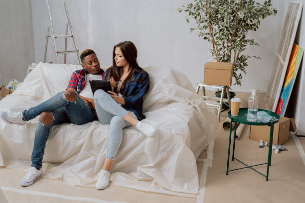 在新买的公寓里，白人女人和深色皮肤的男人坐在客厅的沙发上，装修爱好者们看着平板电脑上的设计，思考着墙壁的装饰颜色图片下载