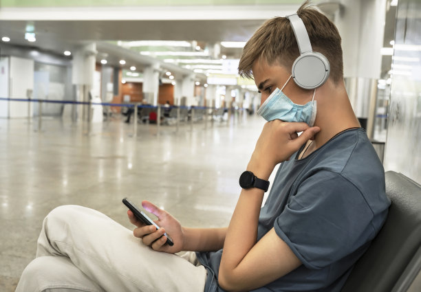 旅客正在等待办理登机手续。15 ~ 18岁的英俊少年戴着防护口罩，在候机室用手机聊天。图片下载