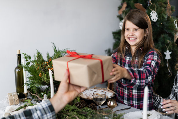 圣诞快乐。快乐的小女孩拿着礼品盒坐在节日桌上。家庭节日的圣诞晚餐在一个装饰的桌子。图片下载